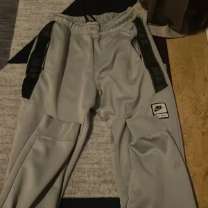 Nike Air Max grå mjukisbyxor S - Grå mjukisbyxor från Nike Air Max med svarta detaljer längs sidorna och dragkedjeförsedda fickor. Byxorna har elastisk midja med snörning och en tydlig Air Max-logga på benet. Perfekta för chill eller träning, tillverkade i mjukt syntetmaterial.