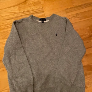 Grå sweatshirt från Polo Ralph Lauren - Klassisk grå sweatshirt från Polo Ralph Lauren med rund halsringning och broderad logga på bröstet. Tröjan har långa ärmar och är tillverkad i mjuk bomullsmix som är perfekt för kyligare dagar. 
