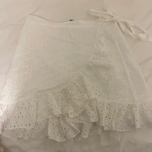 Vit omlottkjol med volang - Supersöt vit omlottkjol med broderade hål och volangkant. Kjolen knyts i sidan och har en luftig känsla, perfekt för varma dagar. 