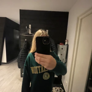 Grön oversized sweatshirt Association - Mörkgrön oversized sweatshirt med gul text 'ASSOCIATION' och ett collegeinspirerat emblem framtill. Tröjan har rund halsringning och långa ärmar, perfekt för en chill och sportig look.