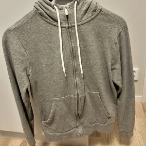 Grå hoodie med dragkedja från Etirel - Grå hoodie från Etirel i storlek S. Tröjan har huva med vita snören, hel dragkedja framtill och två stora fickor. Perfekt för chill dagar eller när du vill ha något bekvämt och enkelt. Mjuk bomullskänsla och klassisk passform.