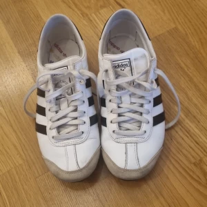 Adidas Originals vita sneakers - Klassiska Adidas Originals sneakers i vitt skinn med svarta ränder på sidorna och svart detalj på hälen. Skorna har snörning, rund tå och platt sula. Insidan har färgglada Adidas-loggor. Perfekta för dig som gillar retrostil och streetwear.