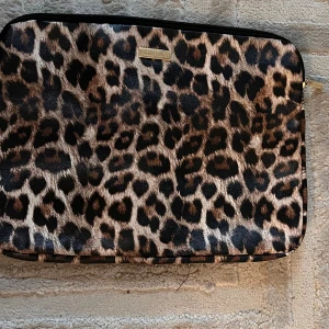 Leopardmönstrad datorväska Cosmopolitan - Snygg datorväska från Cosmopolitan med leopardmönster i bruna och svarta toner. Väskan har dragkedja och är tillverkad i syntetmaterial. Perfekt för dig som vill sticka ut med din tech-stil.