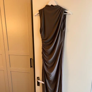 Brun långklänning från Zara i M - Snygg brun långklänning från Zara i storlek M. Klänningen har draperad effekt på sidan och är ärmlös med en elegant, figurnära passform. Tillverkad i ett mjukt och följsamt material som ger ett lyxigt intryck.