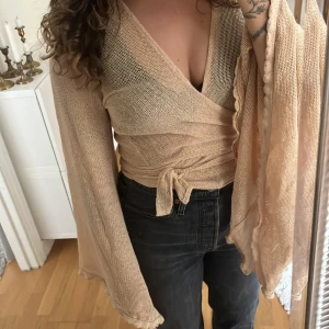 Beige omlottopp från Moonlight - Säljer en superfin beige omlottopp från Moonlight med långa, vida ärmar och knyt framtill. Toppen är i ett tunt, nätliknande material som känns luftigt och snyggt. Perfekt att styla över ett linne eller bikini för en chill vibe.