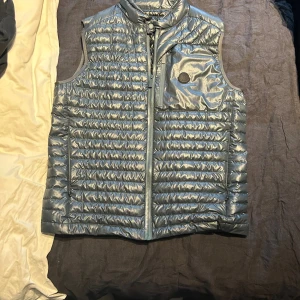 Moncler'Aptera Giubbotto' Gilet Grey - Som helt ny knappt använd. Snygg dunväst från Moncler i metallic silver med quiltad design och hög krage. Västen har dragkedja framtill, bröstficka med dragkedja och diskret Moncler-logga. Perfekt för lager-på-lager och ger en cool vibe till din outfit.