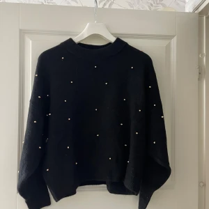 Svart stickad tröja med pärlor - Svart stickad tröja med rund hals och långa ärmar. Tröjan är dekorerad med små pärlor över hela framsidan för en extra snygg touch. Passformen är loose och den har ribbade muddar vid hals, ärmslut och nederkant. Perfekt för dig som gillar stilrena detaljer.
