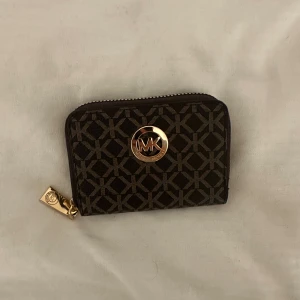 Brun plånbok från Michael Kors - Snygg brun plånbok från Michael Kors med guldiga detaljer och dragkedja runtom. Plånboken har ett grafiskt mönster med MK-logga och flera fack för kort och sedlar. Perfekt storlek för att ha med sig överallt.