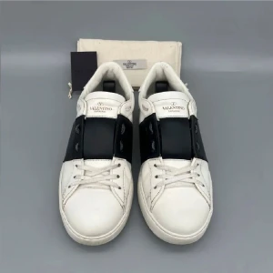 Valentino Garavani Open sneakers vit/svart - Snygga Valentino Garavani Open sneakers i vitt skinn med bred svart läderdetalj över vristen. Klassisk rund tå, vita snören och diskreta nitar på hälen. Perfekta för dig som vill ha en stilren och lyxig look.