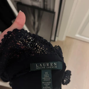 Mörk blå blus med spets från Lauren Ralph Lauren - Säljer en mörk blå blus från Lauren Ralph Lauren i storlek XS. Blusen har lång ärm och en snygg v-ringad hals, som går att knappa upp helt om man vill med dekorativ spets och volangdetaljer. Materialet är mjukt och stretchigt med viskos och elastan för en slim passform. Perfekt för dig som gillar stilrena och feminina detaljer.