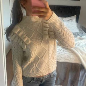 Beige stickad tröja med volanger - Superfin beige stickad tröja med polo och snygga volangdetaljer över axlarna. Tröjan har ett flätstickat mönster på framsida samt långa ärmar. Perfekt för dig som gillar mysiga och trendiga plagg med extra detaljer.