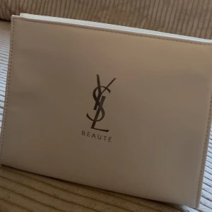 Vit kuvertväska från YSL Beauté - Stilren vit kuvertväska från YSL Beauté med klassisk logga i silver på framsidan. Väskan är tillverkad i syntetmaterial och har ett enkelt magnetlås. Perfekt för att bära det viktigaste och ger en clean look till din outfit. (Äkta och ny)