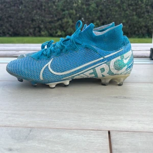 Blå Nike Mercurial fotbollsskor - Säljer ett par blå Nike Mercurial fotbollsskor med vit logga och textdetaljer. Skorna har snörning, högre strumpliknande skaft och plastsula med fasta dobbar. Ovandelen är i syntetmaterial som ger en tight passform och bra bollkontroll. Perfekta för dig som vill sticka ut på planen.