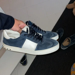 Intressekoll!! Valentino flycrews  - Snygga sneakers från Valentino Garavani i mörkblå mocka med vita skinnpaneler och svarta skosnören. Alla nitar sitter kvar och är i en väldigt fint skick. Klassisk rund tå och platt sula med små nitar på hälen för en extra touch. Perfekta för dig som vill ha en stilren men ändå lyxig look. Kom med prisförslag 