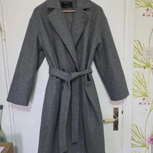 Grå kappa med bälte från Sinsay - Stilren grå kappa från Sinsay med klassisk krage och bälte i midjan. Kappan är helfodrad, har långa ärmar och två fickor framtill. Perfekt för dig som vill ha en tidlös look med snygg passform.