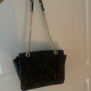 Svart quiltad axelväska med kedja - Snygg svart axelväska med quiltad design och guldfärgad kedjerem. Väskan har ett stort huvudfack, innerficka med dragkedja och magnetlås. Perfekt storlek för det viktigaste och ger en lyxig känsla till din outfit.