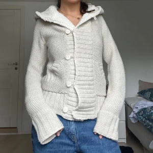 Vit stickad kofta med huva och knappar - Mysig vit stickad kofta med stor huva och grova stickade detaljer. Koftan har stora knappar framtill, ribbade muddar och två fickor. Perfekt för kyliga dagar och har en avslappnad, lite loose passform.
