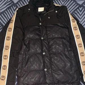 Gucci pufferjacka  - Gucci pufferjacka i svart med diskret GG-mönster över hela jackan. Beigea band med bruna GG-loggor längs ärmarna ger en lyxig vibe. Jackan har knappar framtill och hög krage. Materialet är polyester och jackan är quiltad för extra värme.    Byter mot andra kläder helst jeans. Luvan saknas tyvärr 