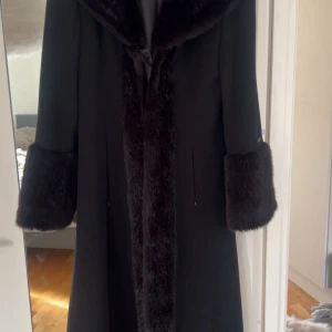 Svart kappa med pälsdetaljer - Säljer en lång svart kappa med bred krage och stora pälsdetaljer vid ärmslut och längs öppningen. Kappan är tillverkad av ull och kashmir, vilket gör den extra mjuk och lyxig. Perfekt för dig som vill ha en elegant och tidlös look under kalla dagar. Strl L men passar mig perfekt som har strl S vanligtvis