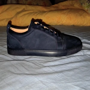 Svarta mockasneakers från Louboutin - Säljer ett par svarta sneakers från Christian Louboutin i mocka med klassisk röd sula och broderad logga på hälen. Skorna har rund tå, platt sula och svarta snören. Insidan är ljusbeige och designen är stilren och lyxig.