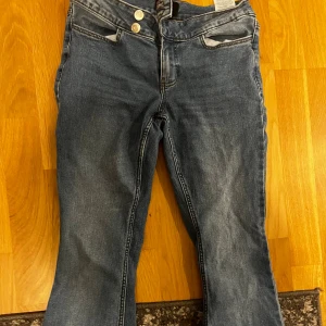 Blå bootcut jeans med dubbla knappar - Säljer ett par blå bootcut jeans med snygg tvätt och dubbla silverfärgade knappar i midjan. Jeansen har klassiska fickor fram och bak, och är tillverkade i mjukt denimtyg. Perfekta för dig som gillar en lite utsvängd look!