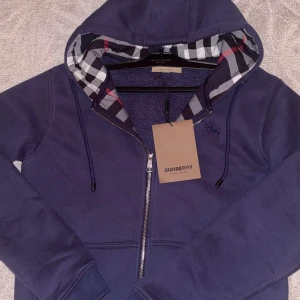 Navy Burberry zip  - Säljer en marinblå hoodie från Burberry med klassiskt rutigt foder i huvan och snyggt broderad logga på bröstet. Tröjan har dragkedja framtill, snörning i huvan och ribbade muddar. Perfekt för dig som vill ha en stilren och lyxig look.