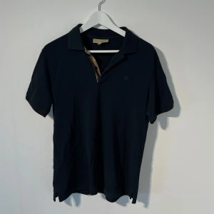 Svart/mörkblå Burberry piké - Svart pikétröja från Burberry med klassisk krage och korta ärmar. Den har en diskret broderad logga på bröstet. Andvänt skick. Storlek L men passar S/M