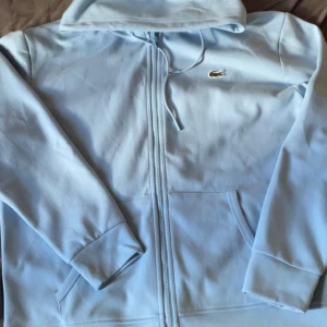 Ljusblå hoodie från Lacoste - Säljer en ljusblå hoodie från Lacoste med dragkedja och huva. Klassisk krokodillogga på bröstet, fickor framtill och snörning i huvan. Skön passform och perfekt för chill dagar. Classic fit-modell.