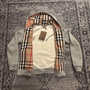 Grå huvud tröja  - Grå hoodie från Burberry med dragkedja, huva och snörning. Insidan av huvan och öppningen har det ikoniska rutmönstret i beige, svart, vitt och rött. Broderad Burberry-logga på bröstet. Mjuk bomullskvalitet och känguruficka framtill.