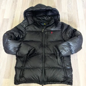 Svart pufferjacka från Polo Ralph Lauren - Svart pufferjacka från Polo Ralph Lauren med huva, dragkedja framtill och flera fickor. Jackan har det klassiska röda Polo-logot broderat på bröstet och är tillverkad i glansig polyester. Perfekt för kalla dagar och riktigt snygg streetstil.