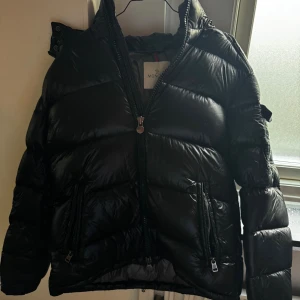 Moncler Maya  - Säljer en svart pufferjacka från Moncler med glansig finish och klassisk logga på ärmen. Jackan har hög krage, två dragkedjefickor framtill och quiltad design. Säljer då den inte passar mig längre. Perfekt för kalla dagar.