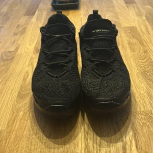 Nike Vapormax svarta sneakers - Svarta Nike Vapormax sneakers med stickad ovandel och cool, genomskinlig sula med blå-lila skimmer. Skorna har snörning och en sportig vibe, perfekt för dig som gillar streetstyle. Swoosh-logga på sidan och Vapormax-detalj på plösen.