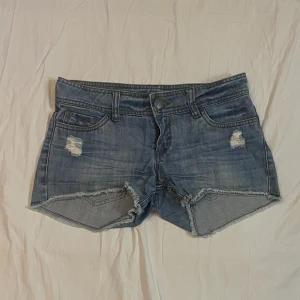 Blå jeansshorts med slitningar - Snygga blå jeansshorts med råa kanter och slitna detaljer framtill. Klassisk femficksmodell med knapp och dragkedja. Perfekta för sommaren och ger en avslappnad vibe.