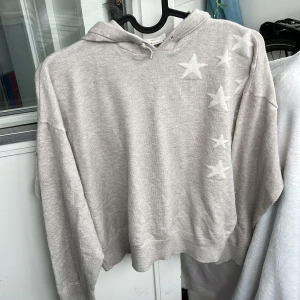 Beige mjuk hoodie med stjärnor från Ginatricot - Beige hoodie med huva och snörning. Tröjan har flera vita stjärnor på framsidan och en stor stjärna på ryggen. Mjuk och skön, perfekt för chill dagar. Långärmad och i ett tunt, stickat material.