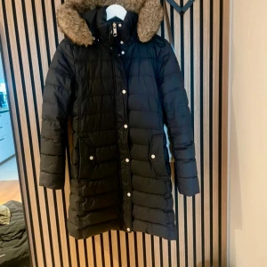 Svart lång dunjacka Tommy Hilfiger - Lång svart dunjacka från Tommy Hilfiger med huva och fuskpäls, quiltad design och dragkedja samt knappar framtill. Jackan har två stora fickor med tryckknappar och logga på ärmen. Perfekt för kalla dagar.