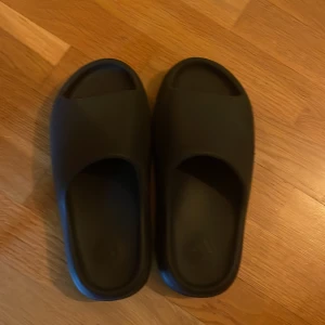 Svarta Adidas Yeezy Slides - Säljer ett par svarta Adidas Yeezy Slides med chunky, ribbad sula och öppen tå. Ny kostar ish 2000kr. De är tillverkade i syntetmaterial och har en clean, minimalistisk look som är riktigt trendig just nu. Perfekta för sommaren eller att chilla hemma.