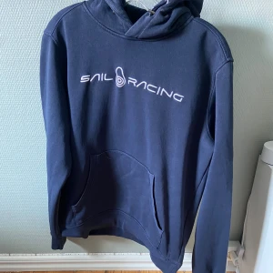 Marinblå hoodie från Sail Racing - Mörkblå hoodie från Sail Racing med vit logga broderad på bröstet. Klassisk känguruficka framtill, huva och långa ärmar. Perfekt för dig som gillar en stilren och sportig look.