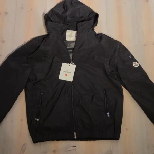 Svart vindjacka från Moncler med huva - Svart vindjacka från Moncler med huva och dragkedja framtill. Jackan har två fickor med dragkedja, elastisk nederkant och klassisk Moncler-logga på ärmen. Perfekt för blåsiga dagar. Snygg och stilren design med diskreta detaljer.