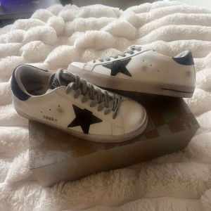Golden Goose sneakers stl 40 - Snygga Golden Goose sneakers i vitt skinn med svart stjärndetalj på sidan och blå hälkappa. Grå snören med logga och klassisk rund tå. Perfekta för dig som gillar en trendig och avslappnad stil. Kommer med originalkartong.