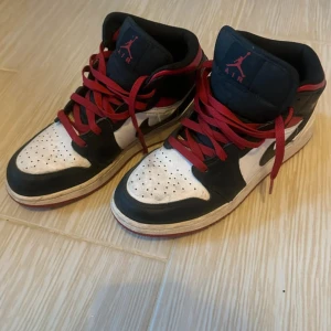 Nike Air Jordan 1 Mid svart/vit/röd 39 - Säljer ett par Nike Air Jordan 1 Mid i klassisk svart, vit och röd färgkombination. Skorna har röda snören, svart och vit ovandel med perforerad tåbox och ikonisk Jumpman-logga på plösen. Tillverkade i skinn och syntet, med rund tå och platt sula.