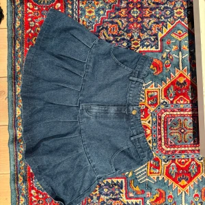 Blå jeanskjol med volang - Säljer en blå jeanskjol med volang och veckad nederdel. Kjolen har klassiska fickor fram och bälteshällor i midjan. Stängs med knapp och dragkedja. Perfekt för dig som gillar en trendig och avslappnad stil.