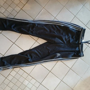 Svarta Adidas mjukisbyxor med ränder - Svarta mjukisbyxor från Adidas med klassiska tre vita ränder längs benen och Adidas-logga på låret. Byxorna har resår i midjan med snörning och dragkedja vid benslut. Perfekta för chill eller träning.