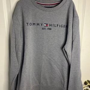 Grå sweatshirt från Tommy Hilfiger - Snygg grå sweatshirt från Tommy Hilfiger med broderad logga och texten 'Est. 1985' på bröstet. Tröjan har rund halsringning, långa ärmar och ribbade muddar. Perfekt för en avslappnad och stilren look. Storlek: 176 