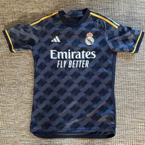 Real Madrid bortatröja Adidas blå - Snygg Real Madrid bortatröja från Adidas i mörkblått med unikt mönster och gula detaljer på ärmarna. Tröjan har klubbmärke och sponsortryck på bröstet samt klassiska Adidas-ränder på axlarna. Tillverkad i lätt och ventilerande material.❗️ÄKTA❗️