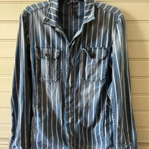 Randig jeansskjorta från Nudie Jeans - Säljer en blå och vit randig jeansskjorta från Nudie Jeans. Skjortan har två bröstfickor med knappar, klassisk krage och långa ärmar. Fet passform och riktigt snygg wash. 