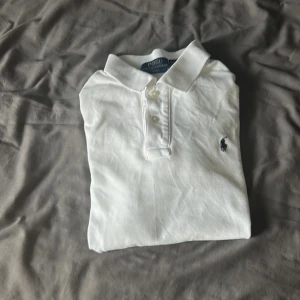 Ralph Lauren pike - Klassisk vit pikétröja från Polo Ralph Lauren med korta ärmar och broderad logga på bröstet. Tröjan har krage, knappar framtill och är gjord i mjuk bomull. Perfekt till jeans eller shorts för en clean och stilren look.