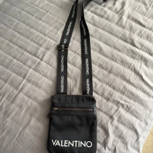 Svart axelväska från Valentino - Snygg svart axelväska från Valentino med justerbart band där loggan står tryckt i vitt. Väskan har en dragkedja framtill.