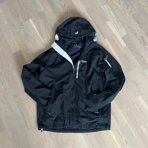 Svart vindjacka från Peak Performance - Svart vindjacka från Peak Performance med huva och dragkedja framtill. Jackan har vita detaljer vid dragkedjor och insida, samt flera praktiska fickor. Materialet är syntet och designen är stilren och sportig, perfekt för blåsiga dagar.