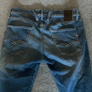 Blå Replay jeans 30/30 - Säljer ett par blå jeans från Replay med snygga kontrastsömmar på bakfickorna och klassisk femficksdesign. Jeansen har en lätt tvättad look och raka ben. Märkesdetaljer på fickorna och midjebandet. Perfekta för dig som gillar stilrena jeans med lite edge.