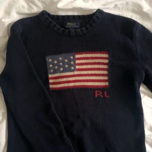 Mörkblå stickad tröja Polo Ralph Lauren - Snygg mörkblå stickad tröja från Polo Ralph Lauren med amerikansk flagga och RL-broderi på bröstet. Klassisk rund halsringning och långa ärmar. Denna är i storlek 14-16 vilket motsvarar L i barn storlek. Kontakta för mer exakta mått. 